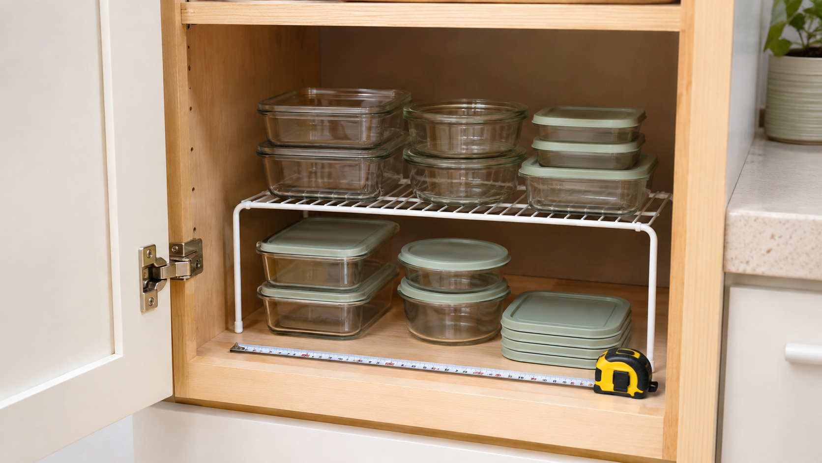 White wire shelf riser separating empty glass meal-prep containers and sage lids inside a wood cabinet, with a measuring tape across the shelf